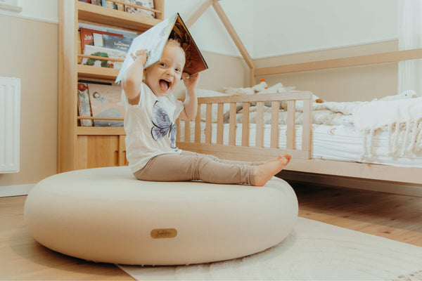Smiling toddler reading on Jupiduu soft pouf in cosy Montessori-style bedroom – playful and calm reading nook with natural wood furniture