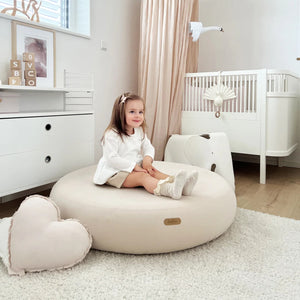 Little girl sitting on a soft and cozy nursery pouf in a stylish, minimalist nursery setup, perfect for kids' playtime and relaxation. Jupiduu Indoor Jumpspot at Little Gliders