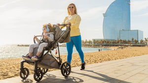 Mother pushing a Mee-go stroller with a child along a sunny beach promenade, featuring a modern skyscraper and palm trees in the background, highlighting comfort and mobility.