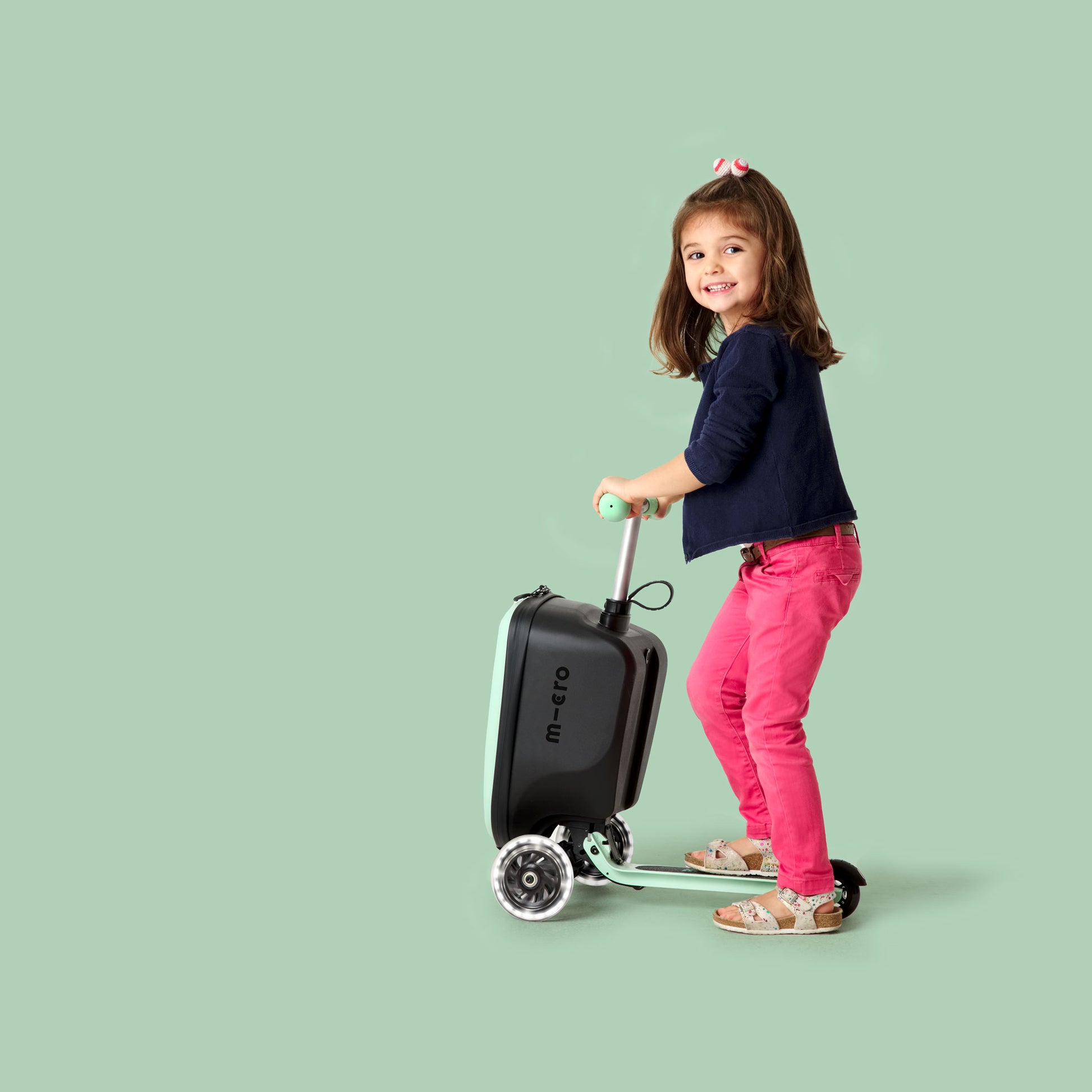 A happy young girl rides a mint green Micro luggage scooter, combining a suitcase with a fun three-wheeled ride. Dressed in pink trousers and a navy top, she smiles confidently against a stylish pastel green background.