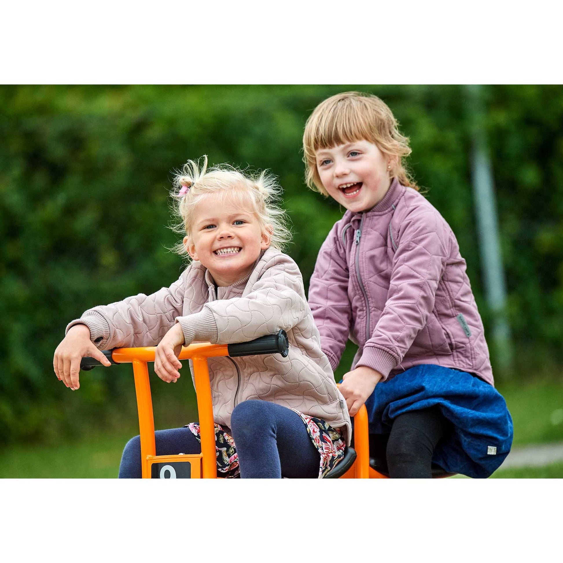 A close-up of two happy girls riding the Winther orange duo trike, enjoying teamwork and outdoor play – available at Little Gliders.