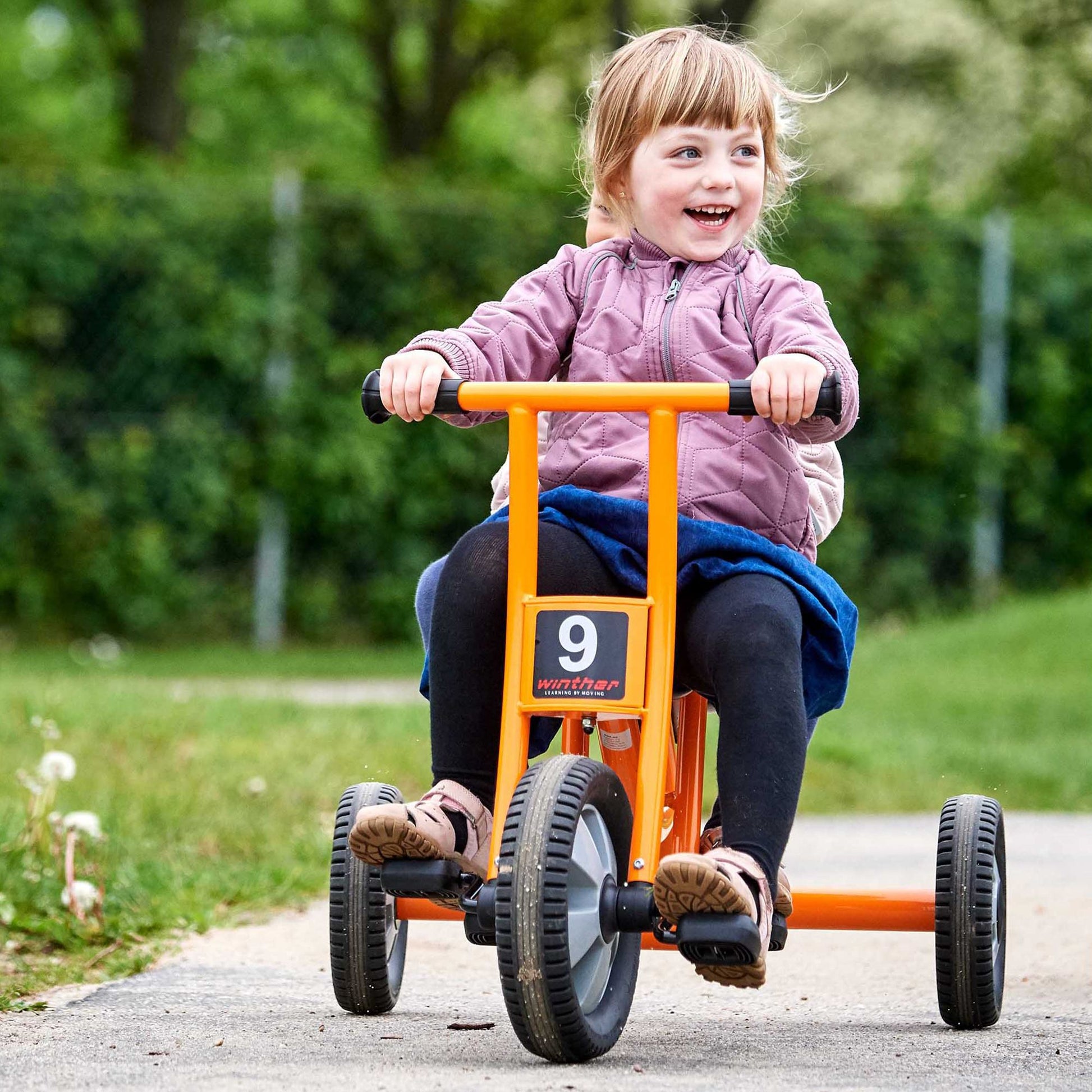 A happy child riding a Winther orange tricycle outdoors, perfect for active play and motor skill development – available at Little Gliders.