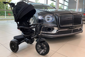 Bentley 6-in-1 Trike in matte black displayed beside a Bentley Flying Spur in a luxury showroom, showcasing its official automotive-inspired design.