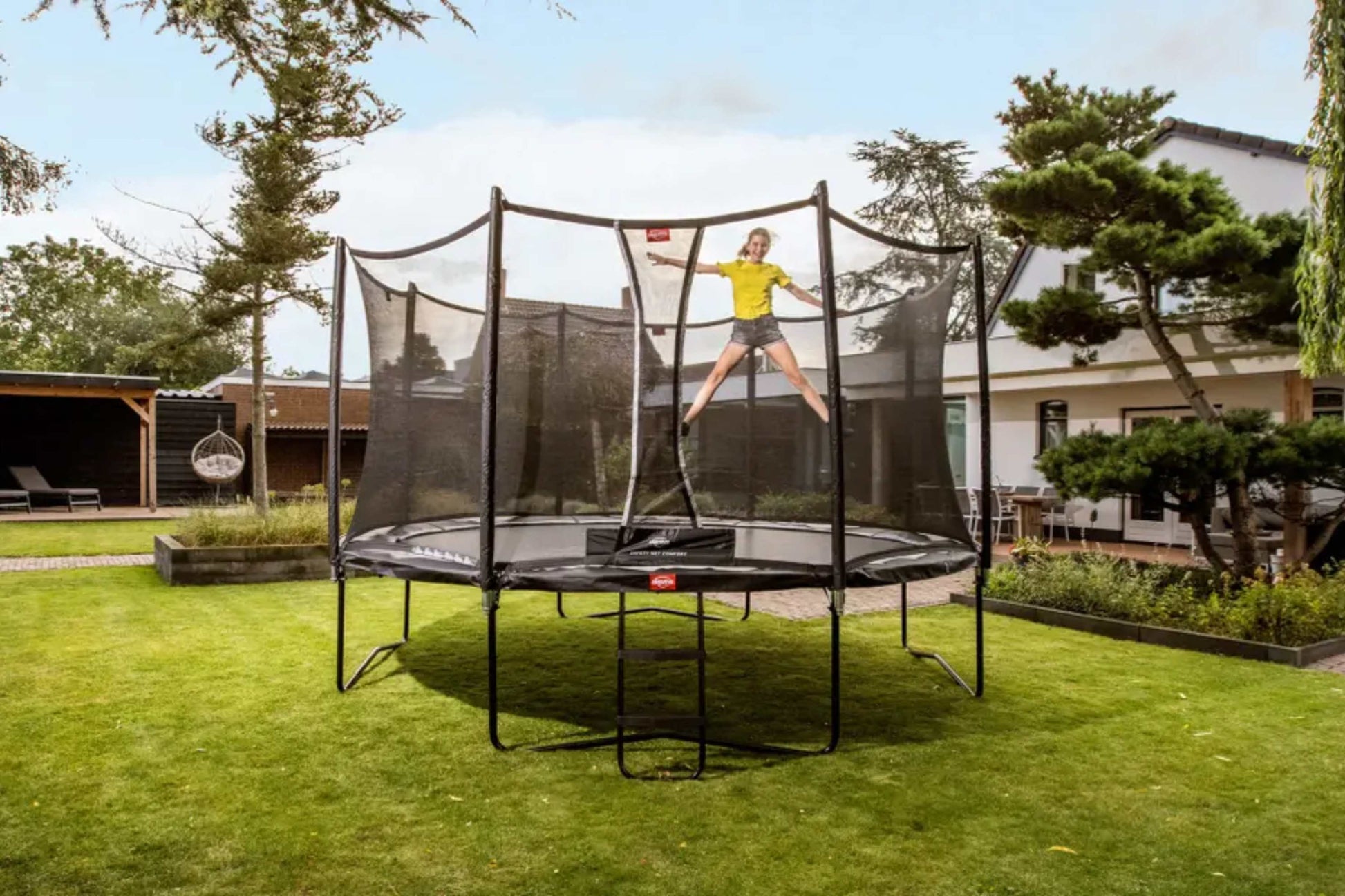 Child jumping on a large round trampoline with safety net enclosure in a modern garden with lawn, ideal for outdoor family play and kids' active fun.