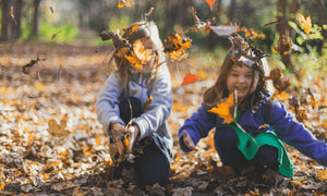 Two young girls playing joyfully with autumn leaves in a forest – perfect for parents searching for children's outdoor clothing, playtime gear, and eco-friendly toys for nature adventures.