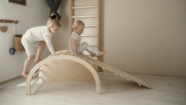 Two toddlers playing on indoor wooden climbing arch with slide, encouraging balance, coordination, and active play in a modern nursery.