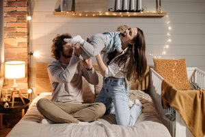 Young parents playing joyfully with toddler on a cosy bed in a stylish nursery with cot, soft lighting, and Little Gliders toys and sleep essentials in the background