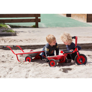 Two young children playing outdoors with red tricycle and wheelbarrow in a sandy play area, encouraging cooperative play, balance, and motor skills.