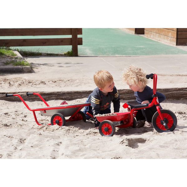 Two young children playing outdoors with red tricycle and wheelbarrow in a sandy play area, encouraging cooperative play, balance, and motor skills.