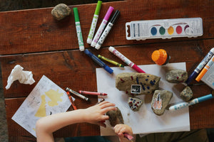 A child creating artwork on rocks using markers and crayons at a wooden desk—symbolising creativity, focus, and the importance of child-sized workspaces.