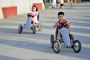 Two smiling children racing outdoor pedal go-karts on a sunny day, building confidence, coordination, and outdoor skills.