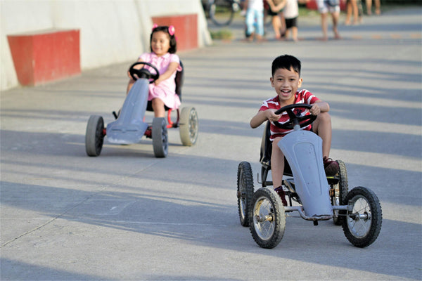 Two smiling children racing outdoor pedal go-karts on a sunny day, building confidence, coordination, and outdoor skills.