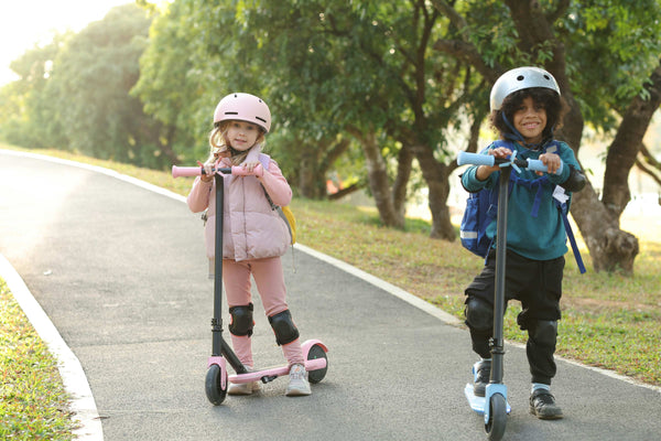 Two young children smiling while riding scooters on a smooth path in the park, wearing helmets and knee pads