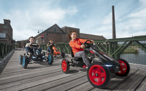 Kids enjoying outdoor adventure on BERG go-karts, racing across a wooden bridge with vibrant wheels and a scenic urban backdrop.