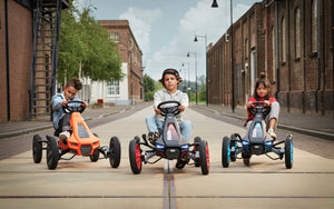 Three children racing side by side on BERG Rally go-karts, featuring vibrant colours and an urban street setting.