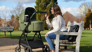 Mother sitting on a park bench, looking at her baby in a green stroller, enjoying a peaceful outdoor moment.