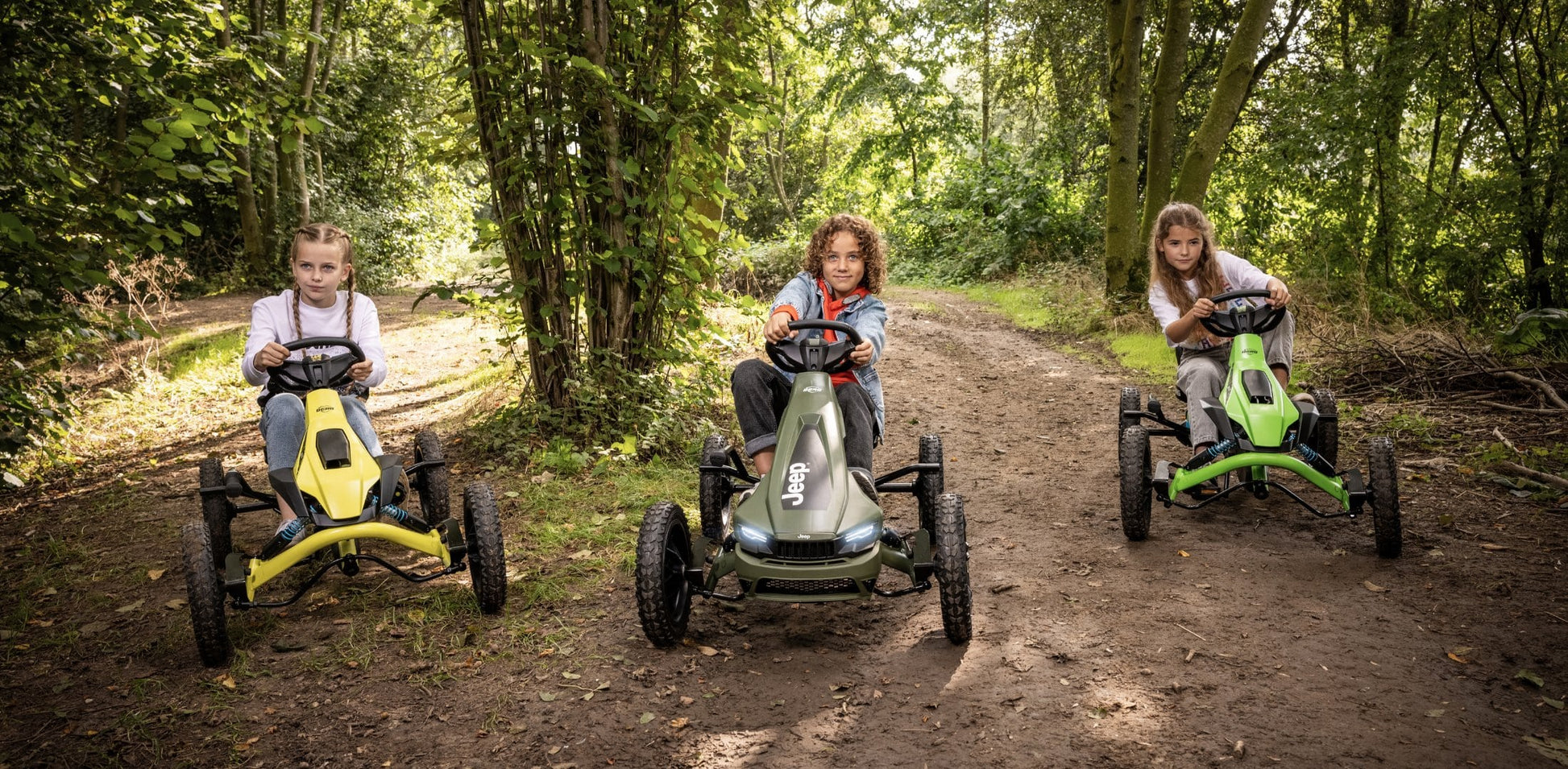 Three kids driving BERG go-karts in a forest trail, featuring yellow, green, and Jeep-themed designs for outdoor adventures