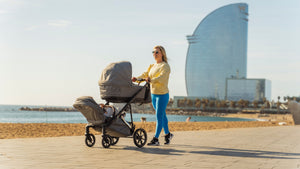 Mother pushing the Mee-go Uno+ 2-in-1 pushchair with two children along a sunny beach promenade, with a modern cityscape in the background.