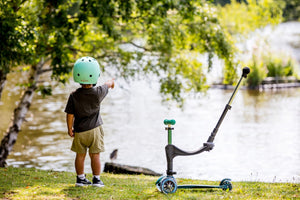 A toddler wearing a mint-green helmet stands near a lake, pointing at the water while their Micro Mini2Grow 3-in-1 scooter rests beside them.
