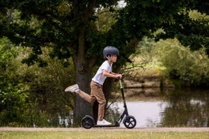 A young child wearing a helmet rides a black Micro Scooter along a scenic riverside path, kicking off with one foot for a smooth glide. Surrounded by lush greenery, the image captures outdoor excitement and active play.