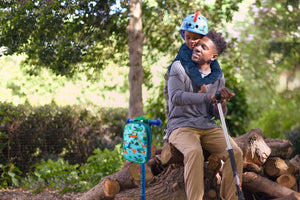 A father and son enjoy a bonding moment in nature, sitting on logs with a Micro scooter and a colourful dinosaur-themed backpack standing beside them.