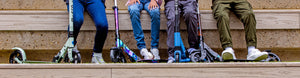  close-up of four kids sitting on wooden steps with their Micro Scooters, showcasing a variety of sleek and colourful designs, built for fun and adventure.