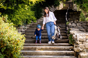 A mother and toddler walk down stone steps outdoors, with the mother carrying a folded Micro Trike 5-in-1, highlighting its lightweight and portable design for families on the go.