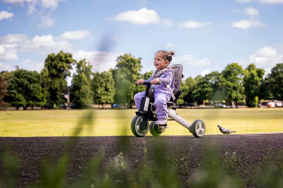 A joyful toddler rides the Micro Trike Deluxe in a scenic park, enjoying a smooth ride in a secure, cushioned seat. The versatile trike features an adjustable design, making it a perfect companion for active little explorers.