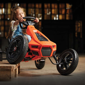 Child playing on an orange BERG go-kart with sleek design and sturdy black wheels in an indoor space.