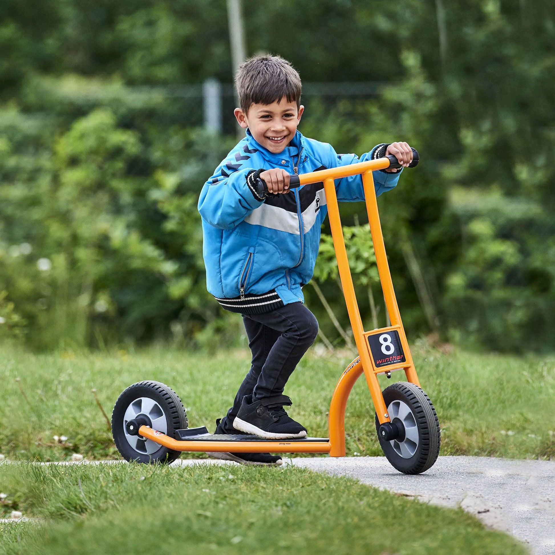 A smiling boy riding a Winther orange scooter outdoors, promoting balance, coordination, and active play – available at Little Gliders.