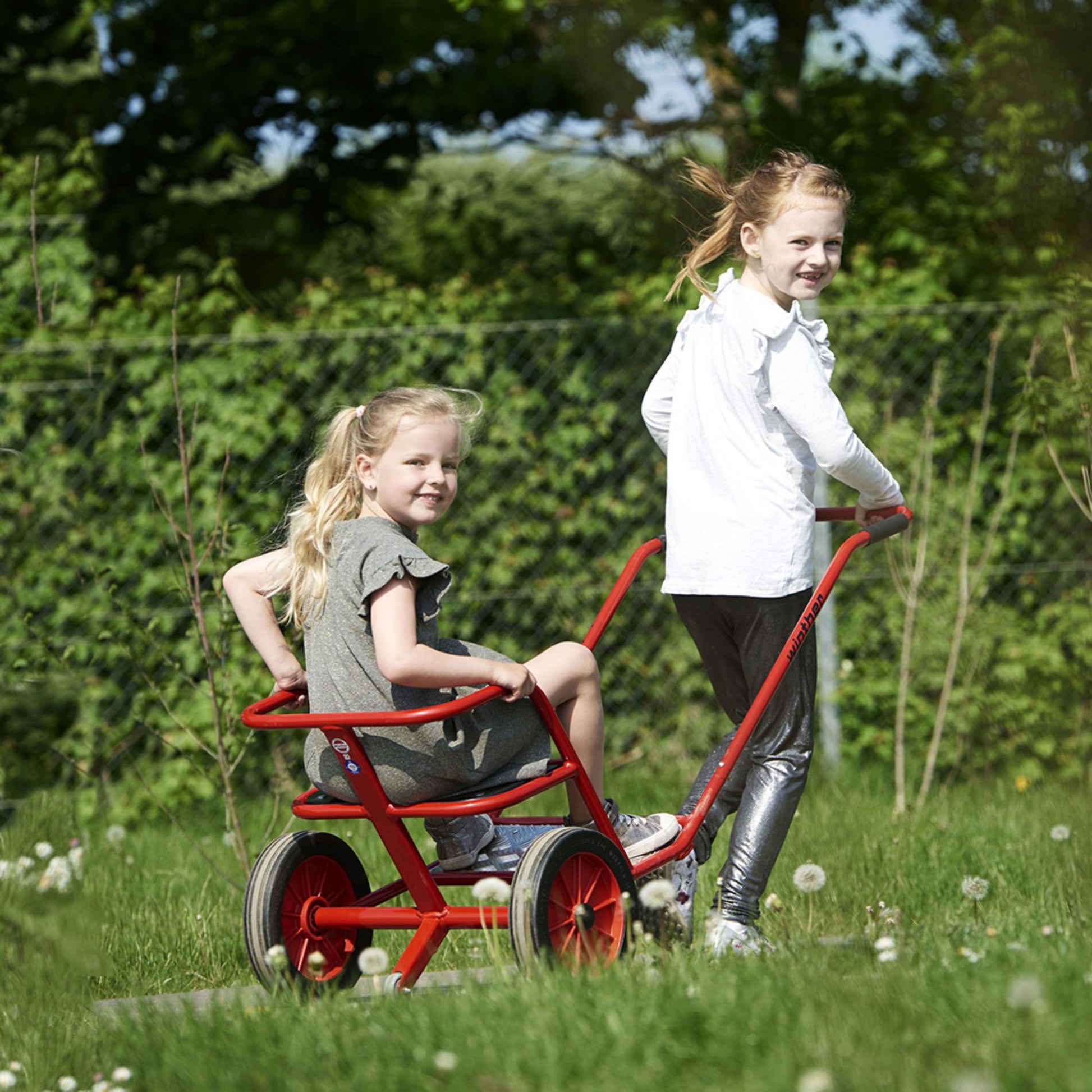 Two girls enjoying a fun ride on a red Winther push-along tricycle, promoting outdoor play and collaboration – available at Little Gliders.